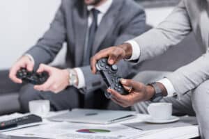 Dois profissionais de terno seguram controles de videogame em uma reunião informal de negócios, com notebook, xícaras de café e documentos sobre a mesa.