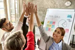Equipe comemorando uma conquista em reunião corporativa, com profissionais levantando as mãos em celebração diante de um quadro com metas e resultados.