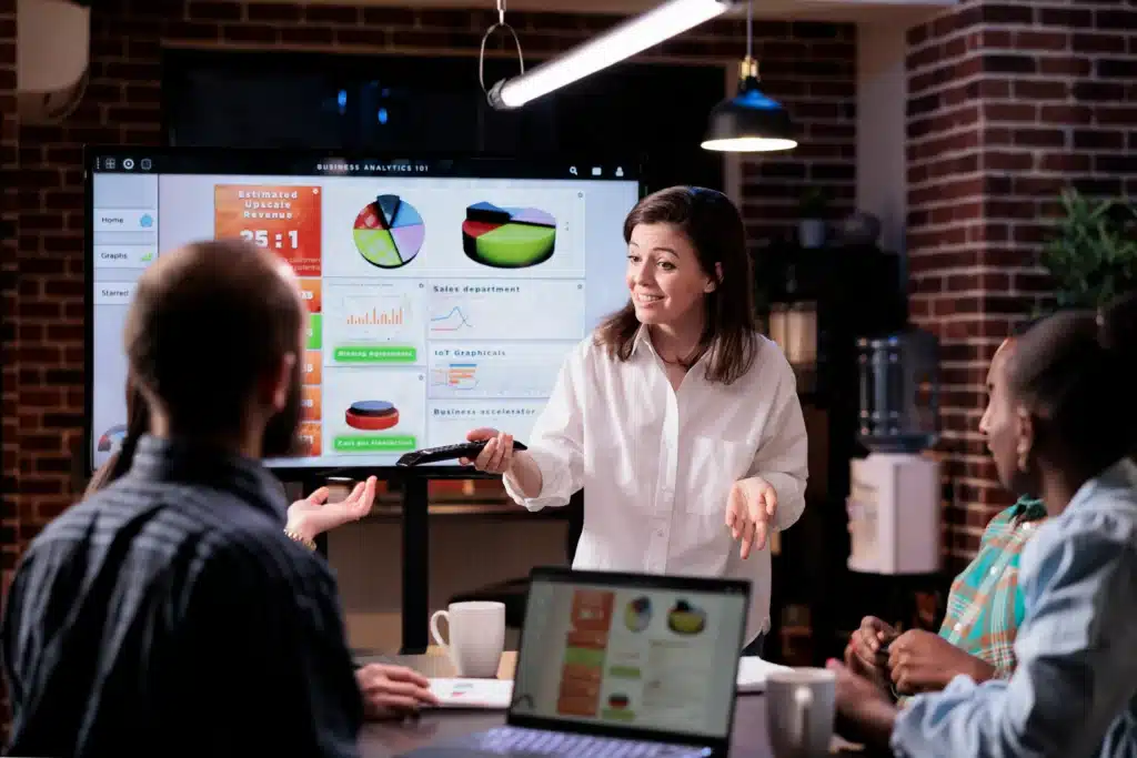 Descrição: Equipe reunida em um ambiente corporativo acompanha uma apresentação sobre resultados e indicadores de desempenho. Uma profissional apresenta informações em um grande painel com gráficos de vendas, receita e performance, enquanto os demais participantes acompanham os dados em uma reunião. A imagem representa análise de resultados, business intelligence, tomada de decisão baseada em dados e acompanhamento de performance.