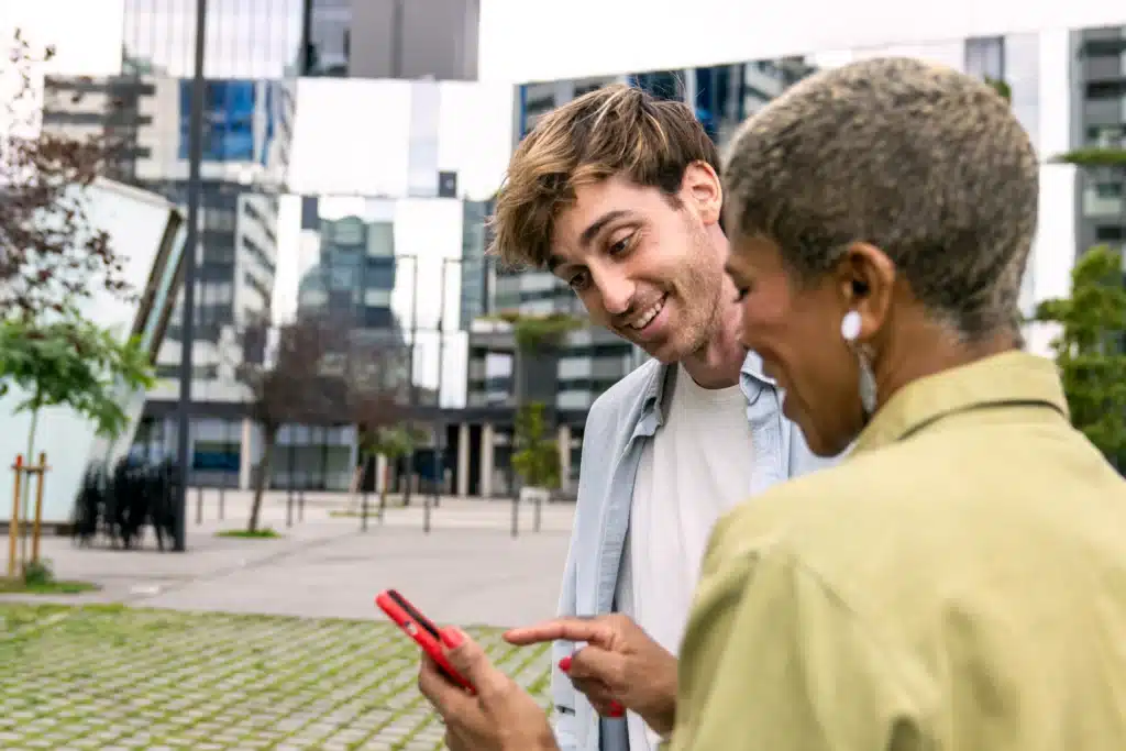 Homem e mulher sorrindo enquanto olham para um celular em uma área externa urbana, com prédios modernos ao fundo.