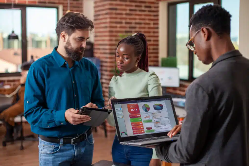 Três profissionais em um escritório moderno analisam indicadores de desempenho utilizando um notebook com gráficos e um tablet. O grupo conversa enquanto avalia dados e resultados, representando colaboração, análise de informações e tomada de decisões estratégicas em equipe.