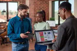 Três profissionais em um escritório moderno analisam indicadores de desempenho utilizando um notebook com gráficos e um tablet. O grupo conversa enquanto avalia dados e resultados, representando colaboração, análise de informações e tomada de decisões estratégicas em equipe.