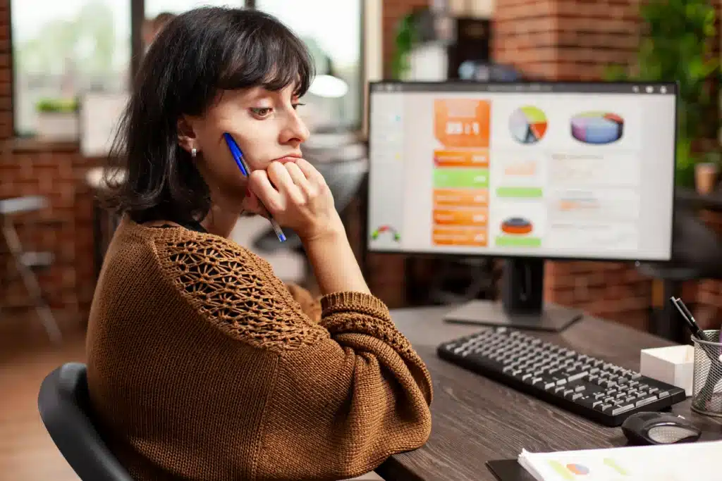 Mulher sentada em uma mesa de escritório observa um monitor com gráficos e indicadores de desempenho enquanto segura uma caneta próxima ao rosto. A expressão pensativa transmite análise, tomada de decisão e avaliação de resultados em um ambiente corporativo moderno.